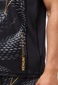 Κοντινό πλάνο μαύρου αθλητικού ρουχισμού με μοτίβο σε γκρι αποχρώσεις που μοιάζει με κλίμακα και κίτρινο κείμενο "VENUM PERFORMANCE" στο πλάι κοντά στο χέρι ενός ατόμου.