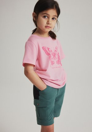 Jong meisje met donker haar draagt een roze t-shirt met vlinderprint en groene cargoshorts met handen in de zakken tegen een effen achtergrond.