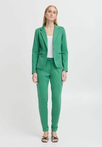 Groene blazer met bijpassende jogger-stijl broek, gemaakt van zachte stof. De blazer heeft een enkele knoopsluiting en voorzakken.