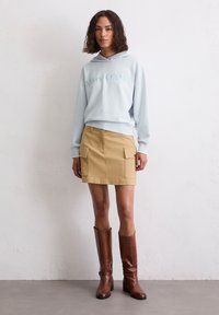 Lichtblauwe hoodie met reliëfletters, gecombineerd met een beige rok met twee grote zakken, aangevuld met bruine kniehoge laarzen.
