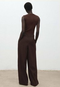 Un adulte avec les cheveux courts portant un haut sans manches marron et un pantalon large marron, se tenant les mains dans les poches contre un mur blanc uni.