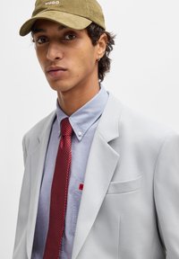 Blazer grigio chiaro su una camicia azzurra chiara, con una cravatta a pois rossi. Gli accessori includono un berretto verde oliva e un orecchino.