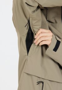 Beige jasstof met een gladde textuur; voorzien van een zijzak met rits en klittenbandaccenten; flexibele ontwerp met een aangepaste pasvorm rond de romp.