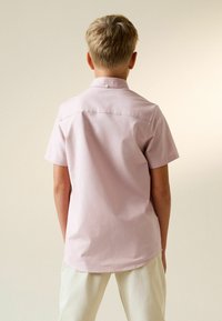 Šviesiaplaukis asmuo dėvi šviesiai rožinę trumpomis rankovėmis marškinių ir baltas kelnes, stovintis nugara į kamerą neutralios spalvos fone.