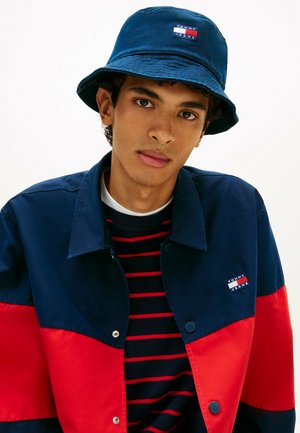 Joven con chaqueta azul marino y roja, sombrero de pescador azul marino y camiseta negra con rayas rojas, mirando directamente a la cámara.