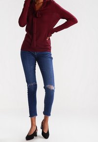 Burgundské triko s dlouhým rukávem a uvazováním na krku, zkombinované s úzkými modrými džíny s roztrhanými detaily a třásněmi na koncích, doplněné černými podpatky.