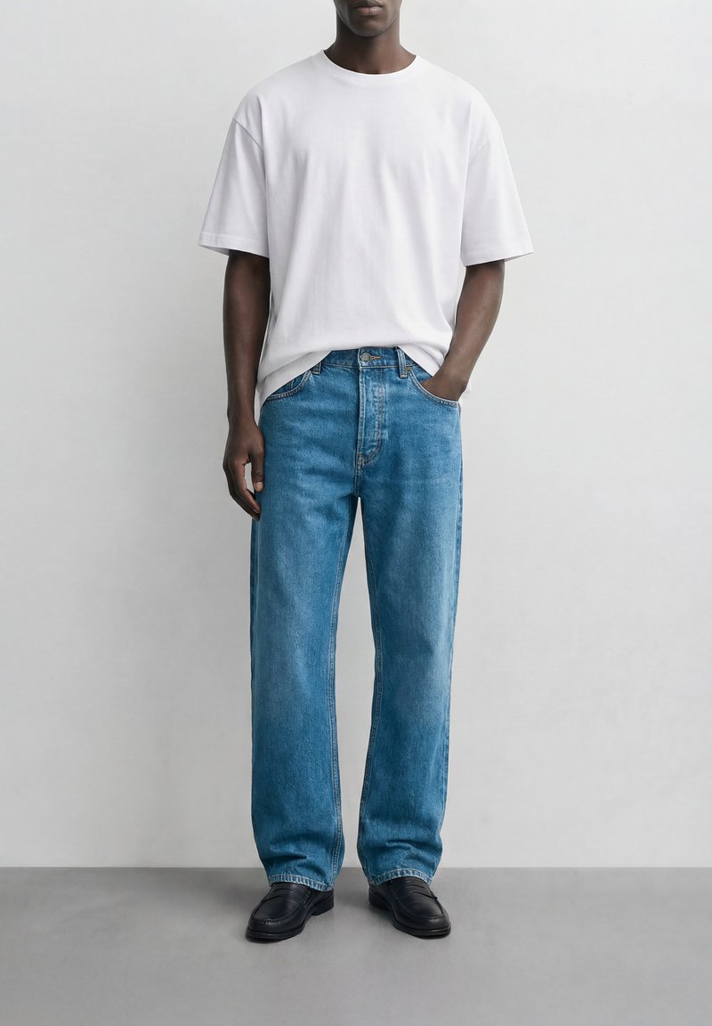 Άνδρας που φοράει φαρδιά μπλε τζιν παντελόνια, μαύρα παπούτσια και απλό λευκό κοντομάνικο μπλουζάκι, στέκεται μπροστά σε ανοιχτό γκρι τοίχο.