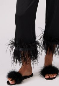 Czarne satynowe spodnie z długimi, piórkowymi wykończeniami w połączeniu z czarnymi futrzanymi klapkami. Posiadają płaską podeszwę i otwarty czubek.