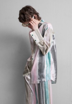 Jeune personne ajustant des lunettes de soleil blanches, portant une chemise ample rayée aux tons pastel et un pantalon assorti, devant un mur gris clair uni.