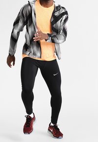 Sportstøj med en mønstret sort og hvid jakke, orange skjorte, sorte leggings og røde sneakers med grå såler.