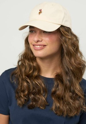 Jauna moteris su ilgais garbanotais rudais plaukais, dėvinti smėlio spalvos beisbolo kepuraitę su arklių logotipu ir tamsiai mėlyną marškinėlį, švelniai šypsosi, žiūrėdama į dešinę.