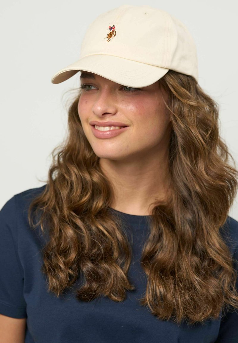 Jeune femme aux longs cheveux bruns bouclés portant une casquette beige avec un logo de cheval et une chemise bleu marine, souriant légèrement et regardant vers la droite.