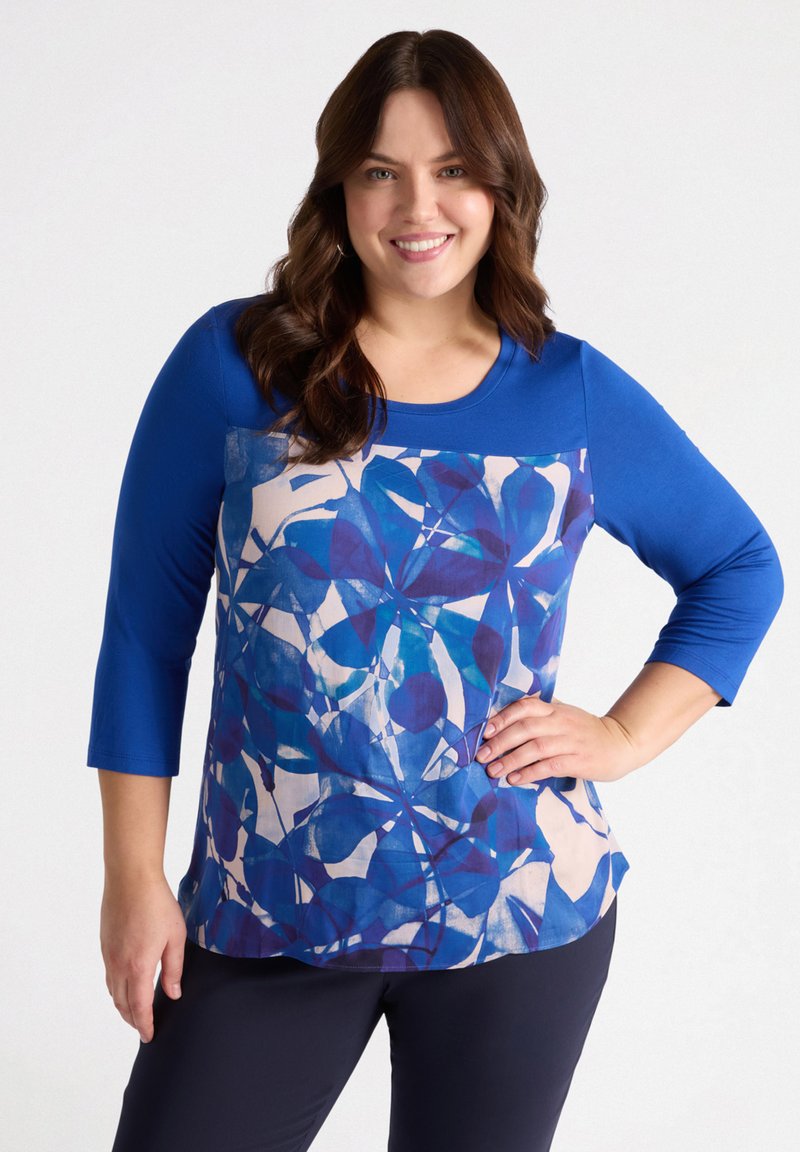 Niebieska bluzka z rękawami o długości trzech czwarte, z kremowym i niebieskim wzorem roślinnym. Posiada okrągły dekolt i gładką teksturę. W zestawie z ciemnymi spodniami.