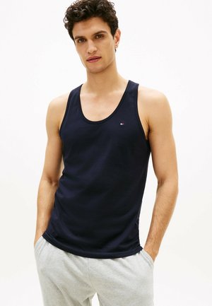 Junger Mann mit dunklem lockigem Haar, der ein marineblaues ärmelloses Tanktop und hellgraue Jogginghosen trägt, Hände in den Taschen, neutraler Hintergrund.