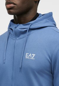 Άνδρας που φοράει μπλε φούτερ με φερμουάρ και λογότυπο EA7 Emporio Armani στο αριστερό στήθος, με εμφανή τον λαιμό και το κάτω μέρος του προσώπου.