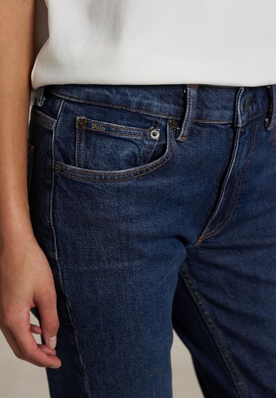 Detailný záber osoby nosiacej tmavomodré denimové džínsy s viditeľnými detailmi predných vreciek a malou štítkom "Polo", v kombinácii s bielym topom.