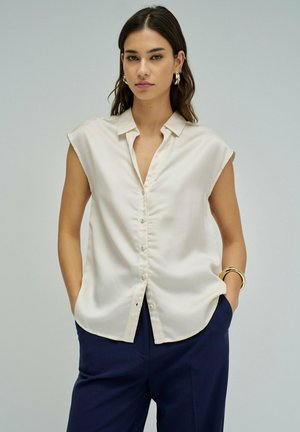 Mujer con cabello largo y oscuro vestida con una blusa sin mangas color crema con botones y pantalones azul marino, complementada con pendientes y pulsera dorados.