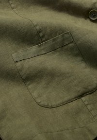 Tela de lino verde oliva con una textura suave; presenta un bolsillo en el pecho y un sutil brillo en el tejido.