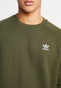 Olivgrön sweatshirt med ribbad rund hals och raglanärmar. Har en liten vit Adidas-logotyp på vänster bröst.
