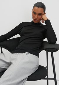 Eine Frau mit dunklem, zurückgebundenem Haar trägt ein schwarzes Langarmoberteil und hellgraue Hosen. Sie sitzt auf einem dunkel gepolsterten Stuhl und stützt ihren Kopf auf die Hand.