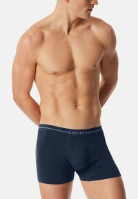 Mörkblå boxertrosor i stretchmaterial med ett märkes elastiskt midjeband med vit text och ribbade detaljer på sidorna.