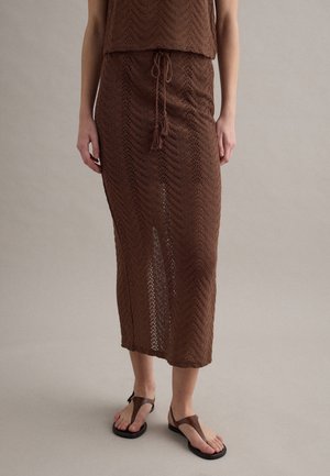 Brauner gestrickter Midirock mit Kordelzugbund, getragen mit passendem Stricktop und braunen flachen Sandalen vor neutralem Hintergrund.