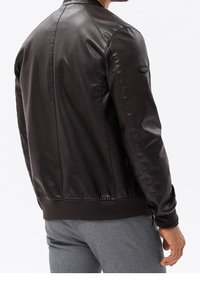 Czarna skórzana bomberka z prążkowanym pasem, charakteryzująca się gładką fakturą i detalem na ramionach. Model ma na sobie szare spodnie.