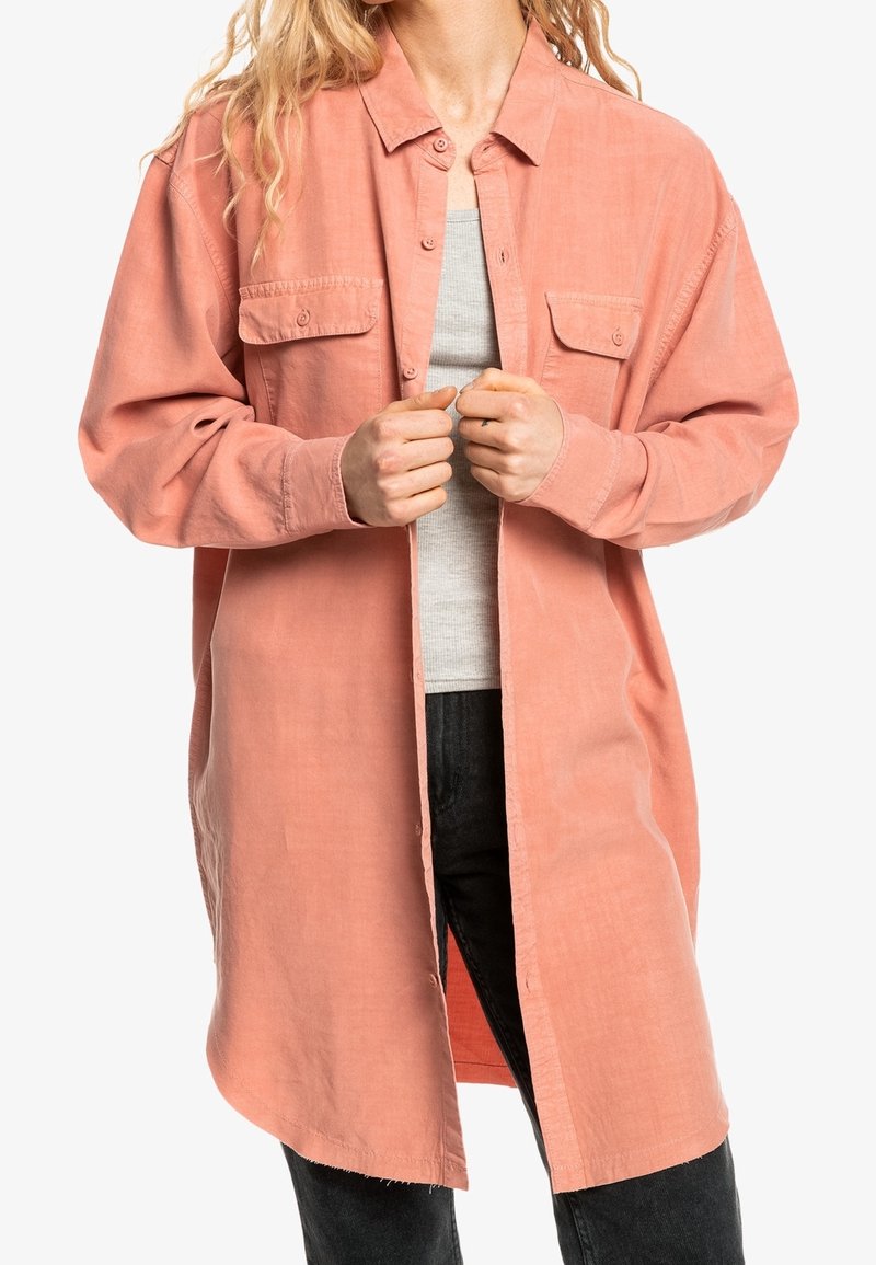 Kobieta zapinająca na guziki oversize'ową koralową koszulę na szary podkoszulek, w czarnych spodniach, widok na tułów, na białym tle.