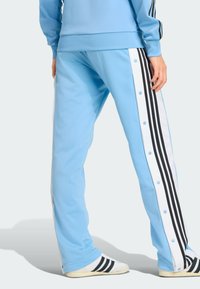Light blue track pants with black stripes, snap-button sides, and a smooth texture. Paired with white shoes featuring black accents.