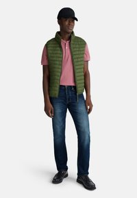 Zielona pikowana kamizelka na różowej koszulce polo, w połączeniu z ciemnoniebieskimi dżinsami i czarnymi loafersami. Czapka jest czarna z logo.