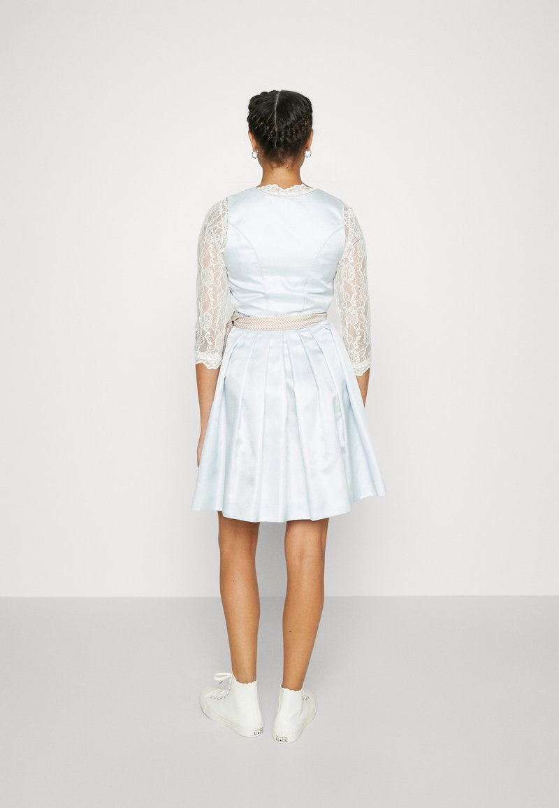 Femme debout de dos à la caméra, vêtue d'une robe plissée bleu clair avec des manches en dentelle et des baskets blanches, sur un fond uni.