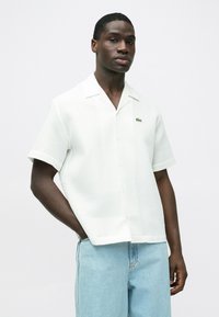 Biela krátka košeľa s textúrovaným vzorom, zapínaním na gombíky a malým zeleným logom Lacoste na hrudi, v kombinácii so svetlomodrými džínsami.