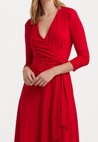 Robe portefeuille rouge en tissu lisse, avec un décolleté en V, des manches longues et un détail de taille nouée, créant une silhouette fluide.