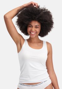 Weißes Baumwoll-Tanktop mit Rundhalsausschnitt, getragen mit passender weißer Unterwäsche mit einem Logo-Bund. Das Model posiert mit erhobener Arm.