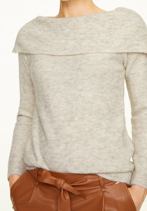 Femme portant un pull gris clair décolleté sur les épaules, rentré dans un pantalon en cuir marron taille haute avec une ceinture nouée, les mains dans les poches.