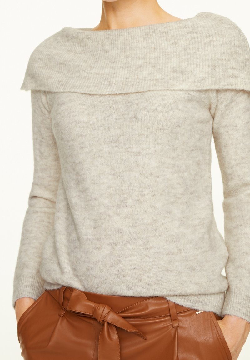 Femme portant un pull gris clair décolleté sur les épaules, rentré dans un pantalon en cuir marron taille haute avec une ceinture nouée, les mains dans les poches.