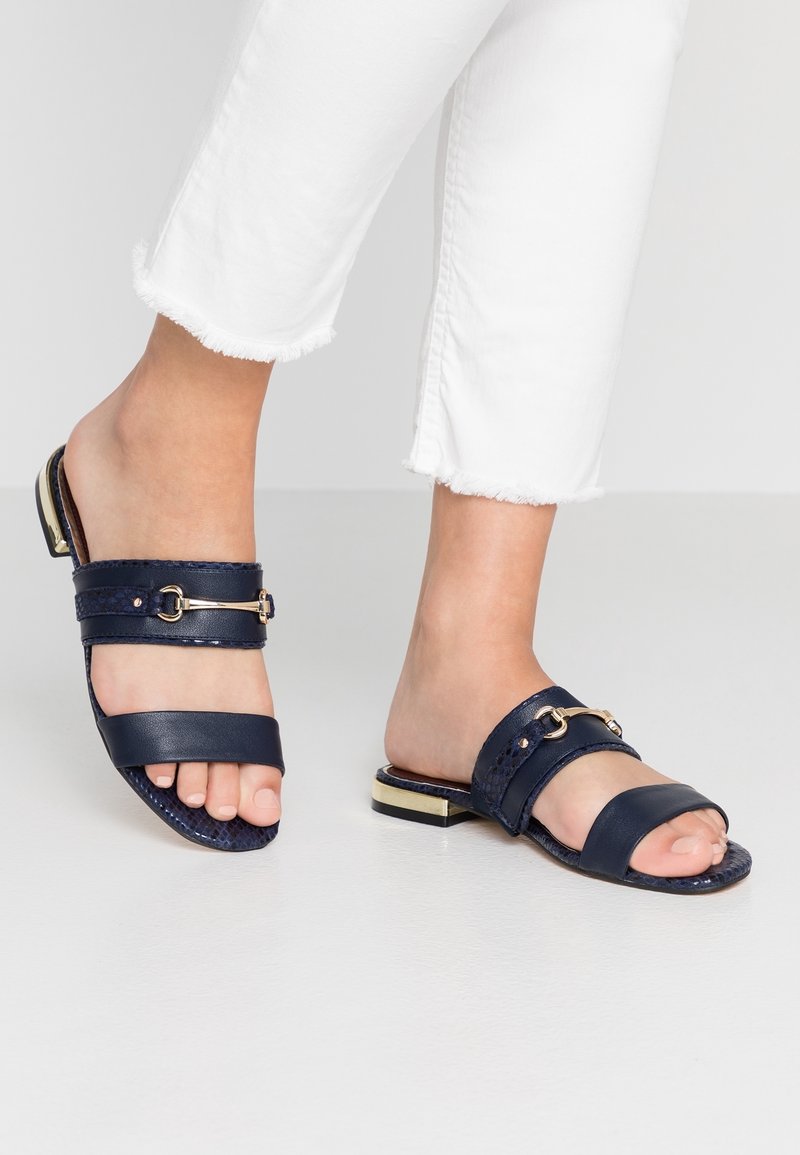 Pieds portant des sandales slides bleu marine avec détail chaîne dorée et talons bas accentués dorés, associés à un jean blanc court effiloché.