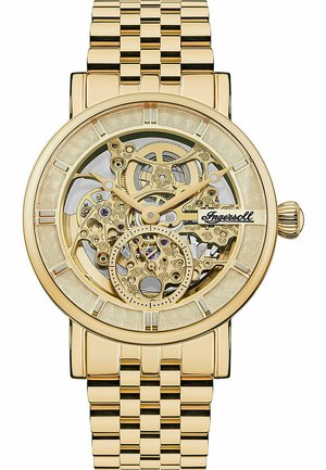 Montre squelette dorée avec un cadran transparent révélant des engrenages complexes. Bracelet à maillons et lunette texturée avec des index horaires.