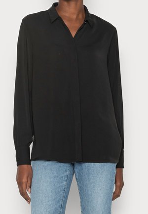 Henkilö, jolla on musta pitkähihainen nappipaita ja vaaleansiniset farkut, seisoo valkoista taustaa vasten.