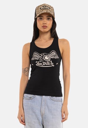 Femme portant une casquette Von Dutch imprimé léopard, un débardeur noir avec un œil ailé et le logo Von Dutch, et un jean bleu clair, debout devant un fond blanc.
