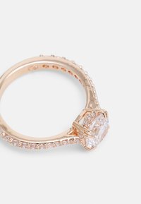 L'anello in oro rosa presenta una pietra centrale trasparente circondata da piccole pietre trasparenti lungo la fascia, con una finitura liscia e lucida.