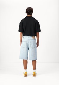 Camisa negra de manga corta con cuello, combinada con pantalones cortos de mezclilla azul claro. Calcetines blancos y zapatillas deportivas con detalles amarillos completan el atuendo.