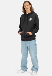 Black hoodie with a front pocket and drawstring hood, featuring a logo on the chest. Paired with light blue, loose-fitting jeans.