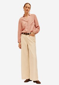 Camicia button-up rosa chiaro con due tasche frontali, abbinata a pantaloni beige a gamba larga e una cintura marrone con fibbia circolare.