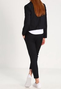 Žena s dlouhými rudými vlasy, oblečená v černém pruhovaném obleku a bílých teniskách, stojící s rukama v kapsách, otočená zády k jednobarevnému pozadí.