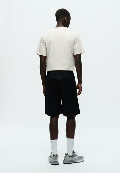 T-shirt beige clair à manches courtes associé à un short noir arrivant au genou. Le mannequin porte des baskets grises et des chaussettes blanches. Design minimal, sans motifs.