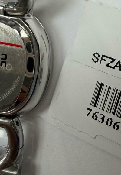 Detailní záběr na zadní část stříbrných hodinek s výrobními čísly vedle bílé etikety s čárovým kódem a kódem "SFZA" na bílém povrchu.