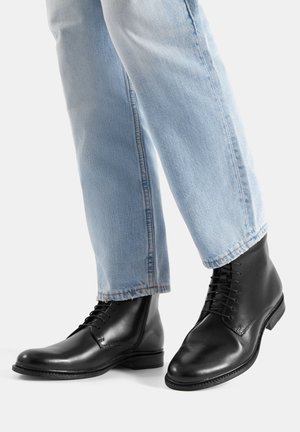 Melnas ādas auklu zābaki ar noapaļotu purngalu un gludu apdari, kopā ar gaiši ziliem taisnas kājas džinsiem, demonstrējot zābaku dizainu un tekstūru.