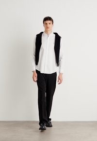 Λευκό πουκάμισο με κουμπιά, φορώντας έναν μαύρο ύφασμα στους ώμους, συνδυασμένο με μαύρα παν trousers και παπούτσια, σε ουδέτερο φόντο.