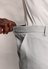 Pantaloni grigi chiari con una finitura testurizzata e una vita piatta, tirati in vita. Primo piano dei dettagli del tessuto e della vestibilità.